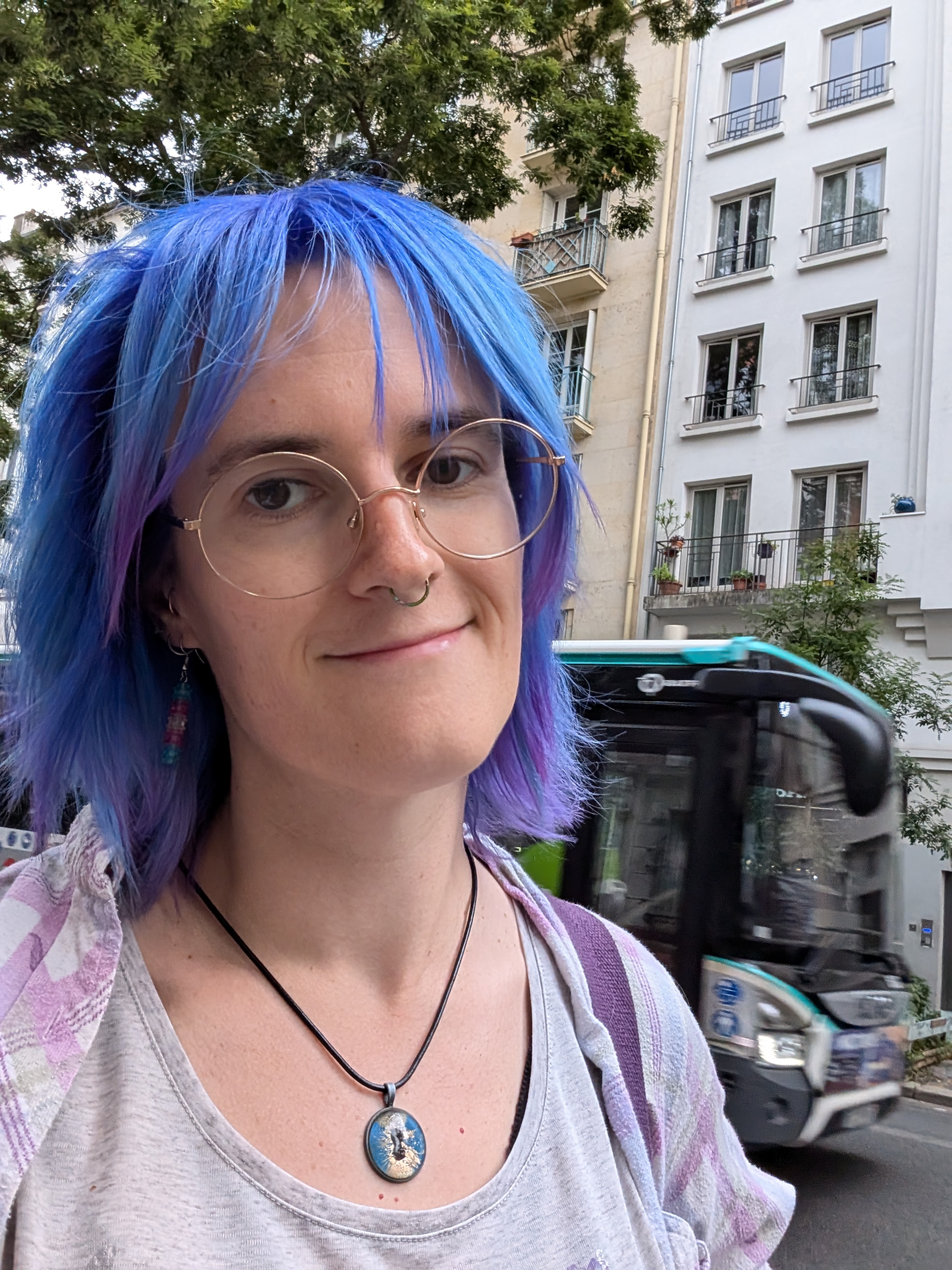 A white woman with blue hair, round glasses and a septum ring is smiling, wearing a white and pink shirt over a gray top.