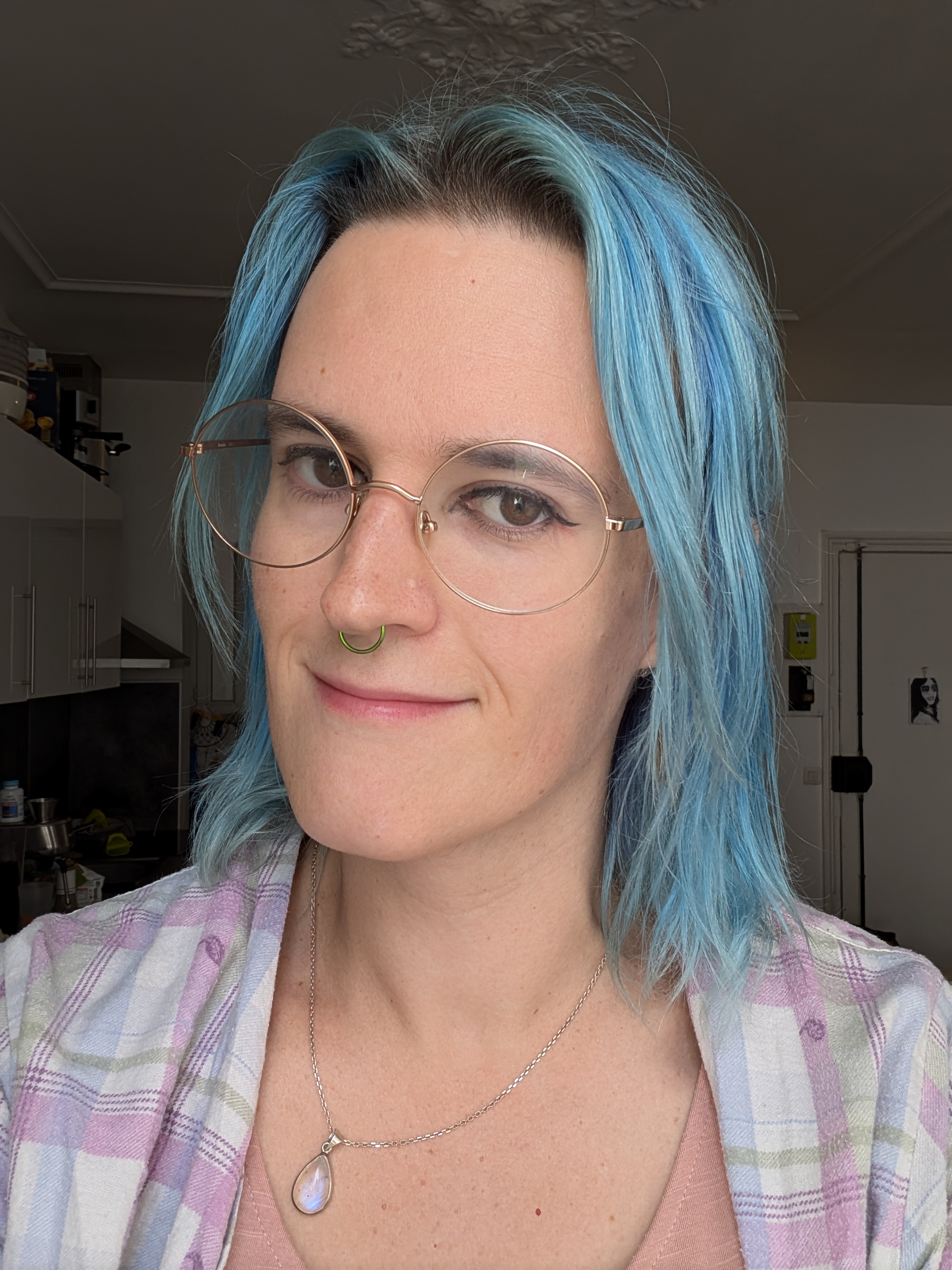 A white woman with blue hair, round glasses and a septum ring is smiling, wearing a white and pink shirt over a pink top.