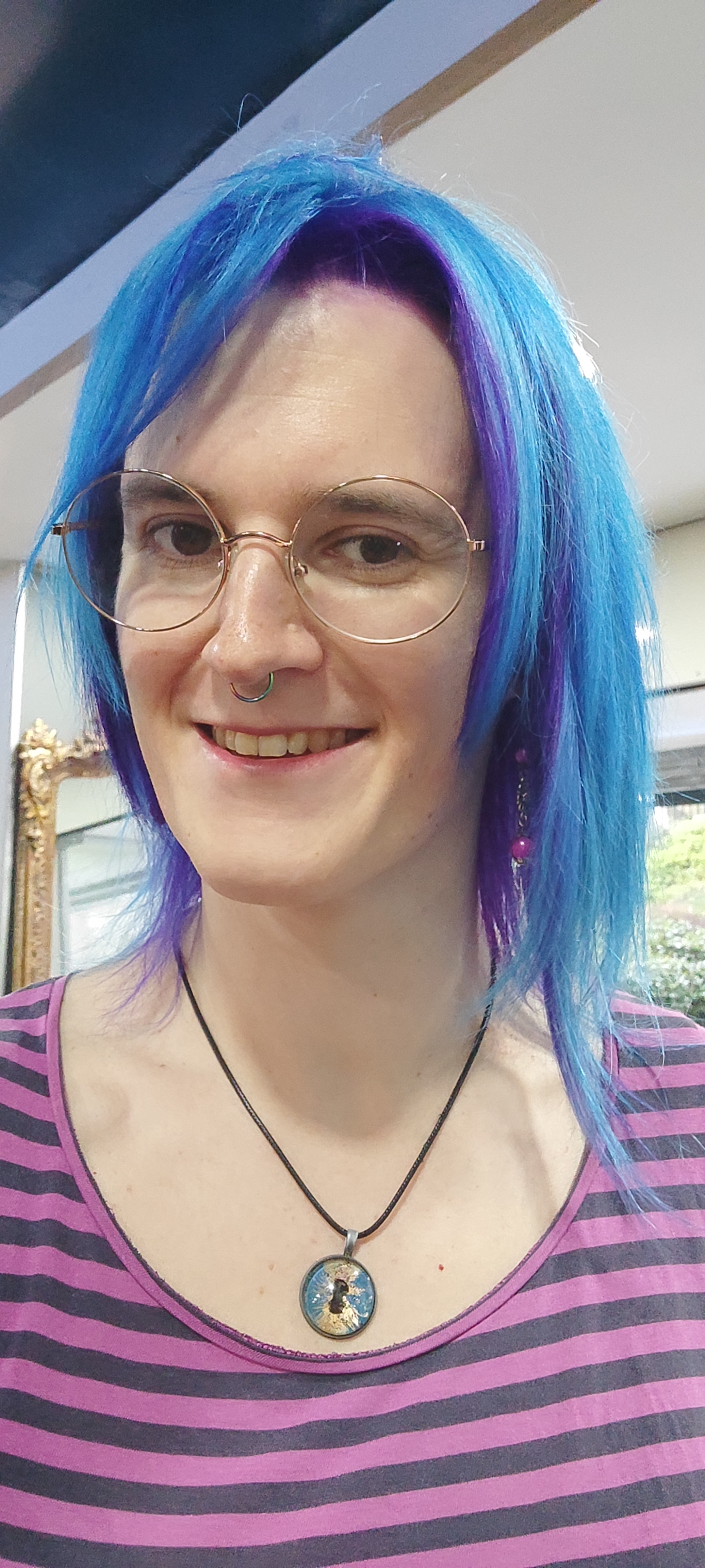 A white woman with blue and purple hair, round glasses and a septum ring is smiling, wearing a striped pink and black top.